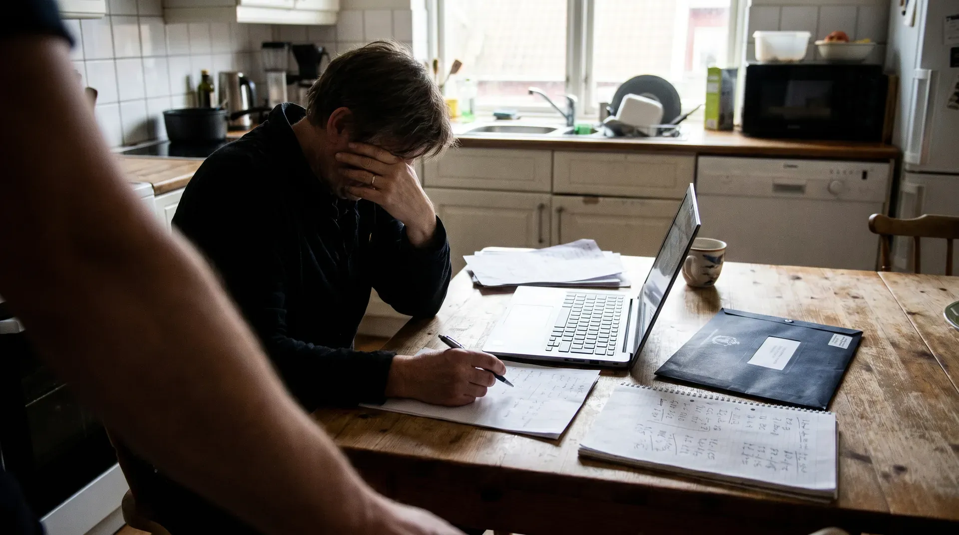 Man aan keukentafel met laptop en papieren, hand aan gezicht in concentratie