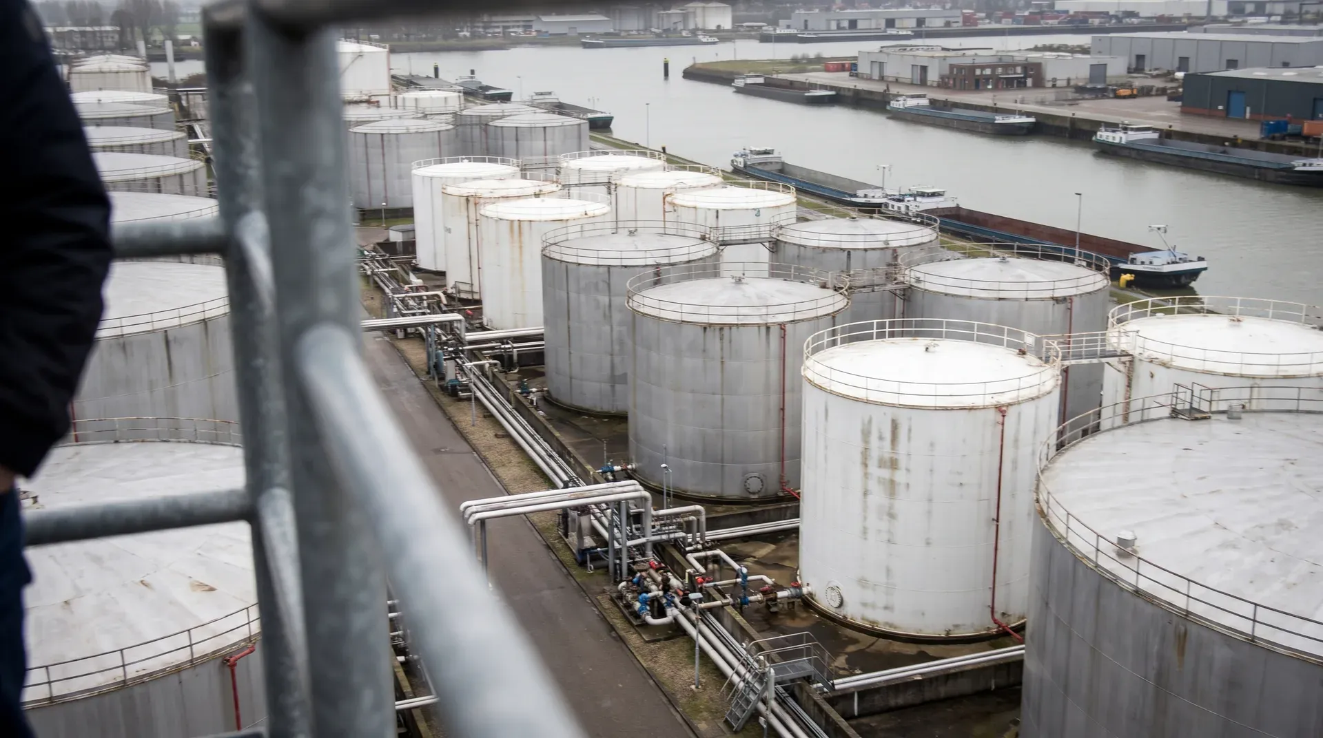 Industriële opslagtanks aan waterfront met binnenschepen en dokgebouwen op achtergrond.