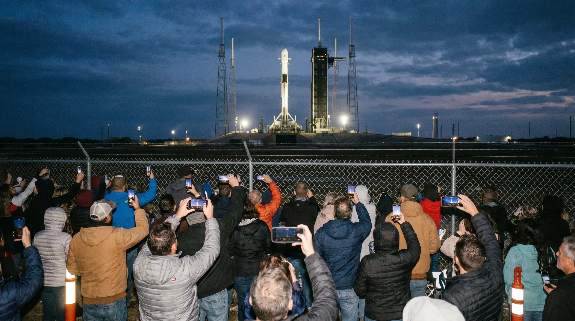 Menigte toeschouwers achter hek fotografeert SpaceX-raket op startplatform bij schemering