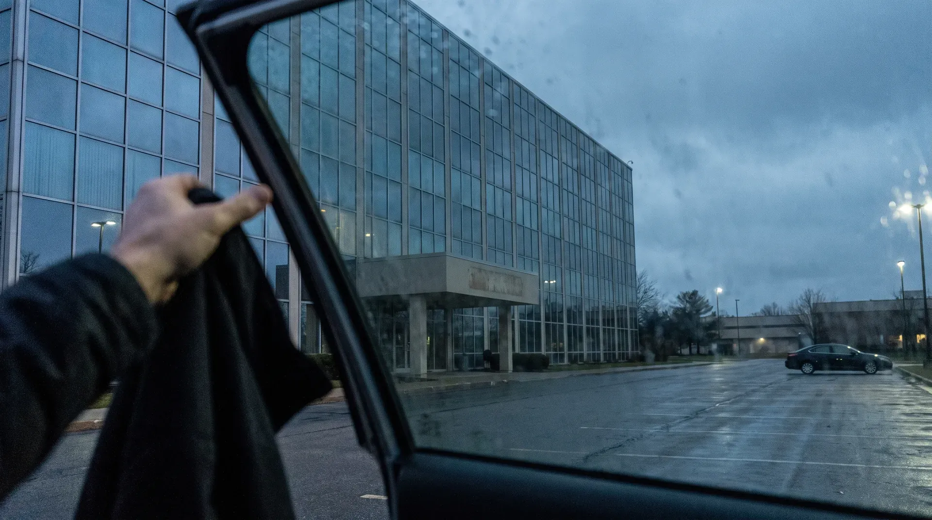 Persoon in auto kijkt naar modern glazen kantoorgebouw in schemering op natte parkeerplaats