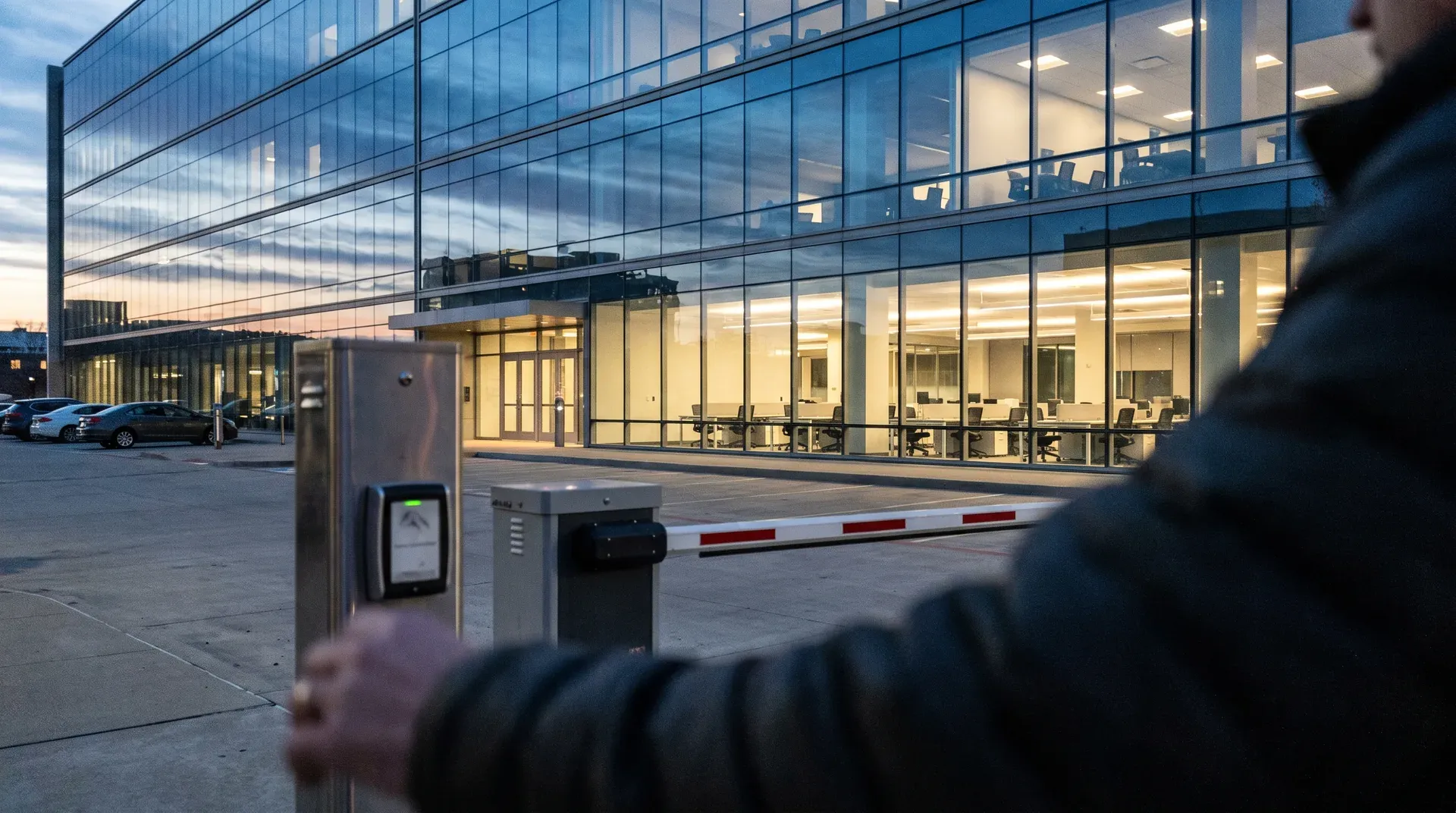 Modern glazen kantoorgebouw bij schemering met verlichte ramen, toegangspoort en parkeerplaatsen.