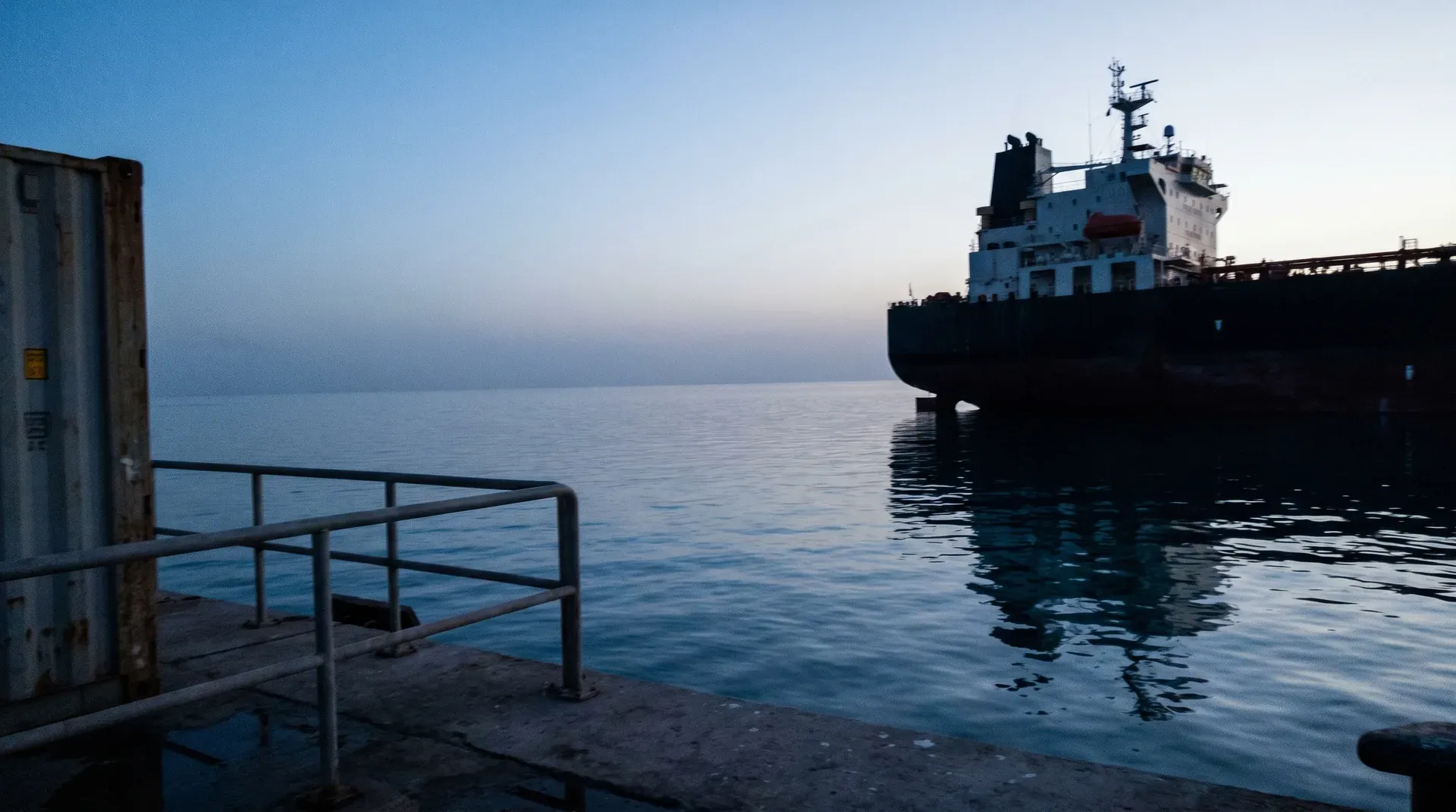 Tanker in kalme zee bij schemering, gezien vanaf industriële havenkaai met railing
