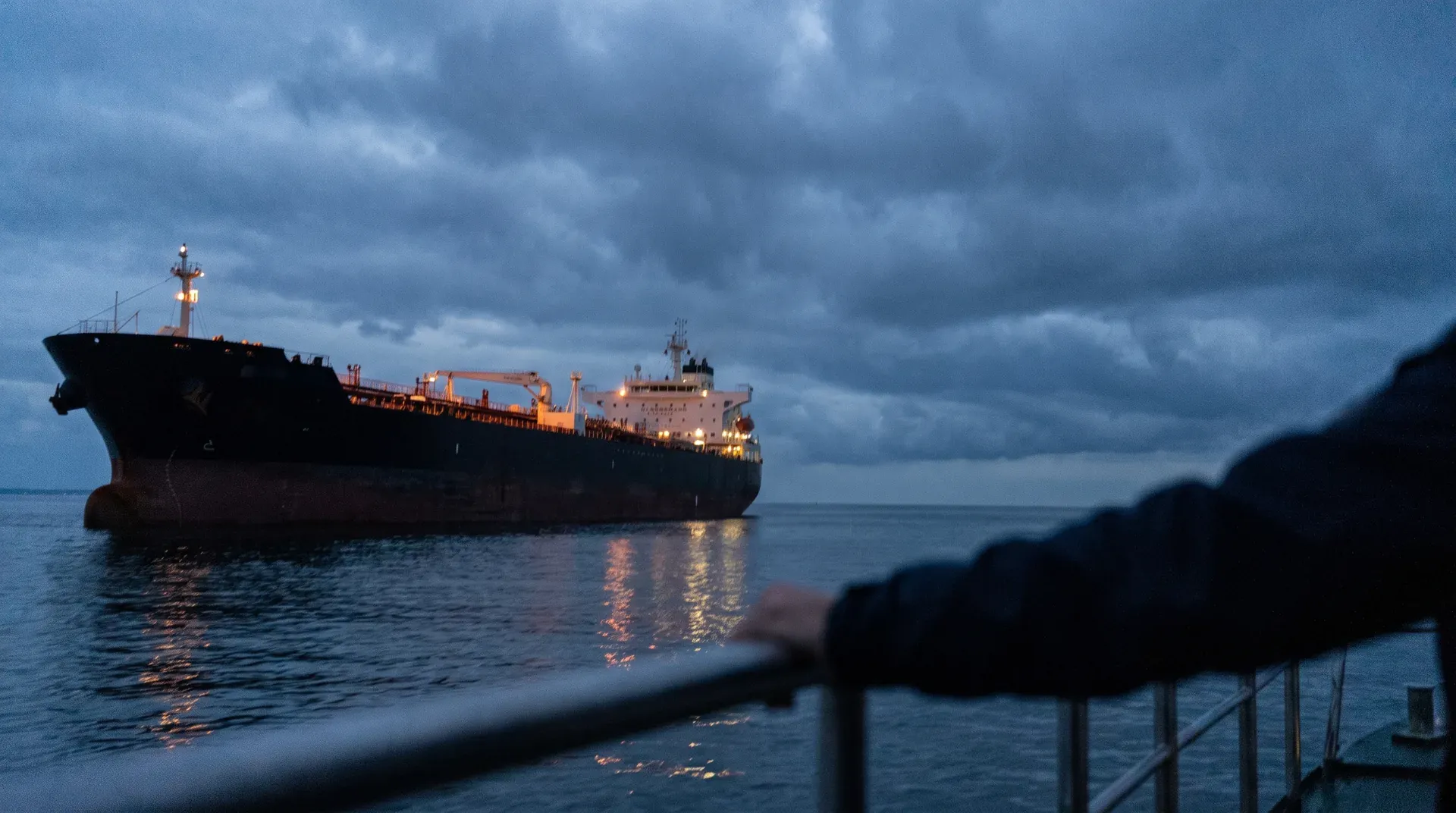 Verlichte olietanker in zee tijdens schemering onder bewolkte hemel, gezien vanaf kust