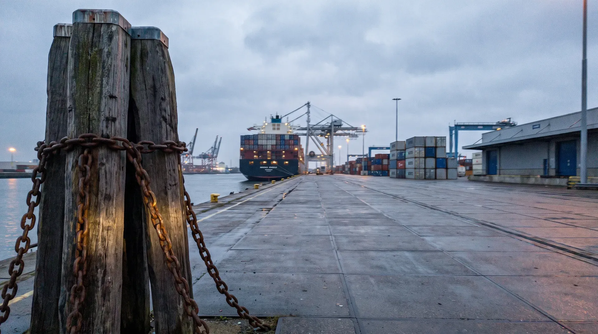 Containerschip bij containerterminal met kranen, roestige paal met ketting op voorgrond