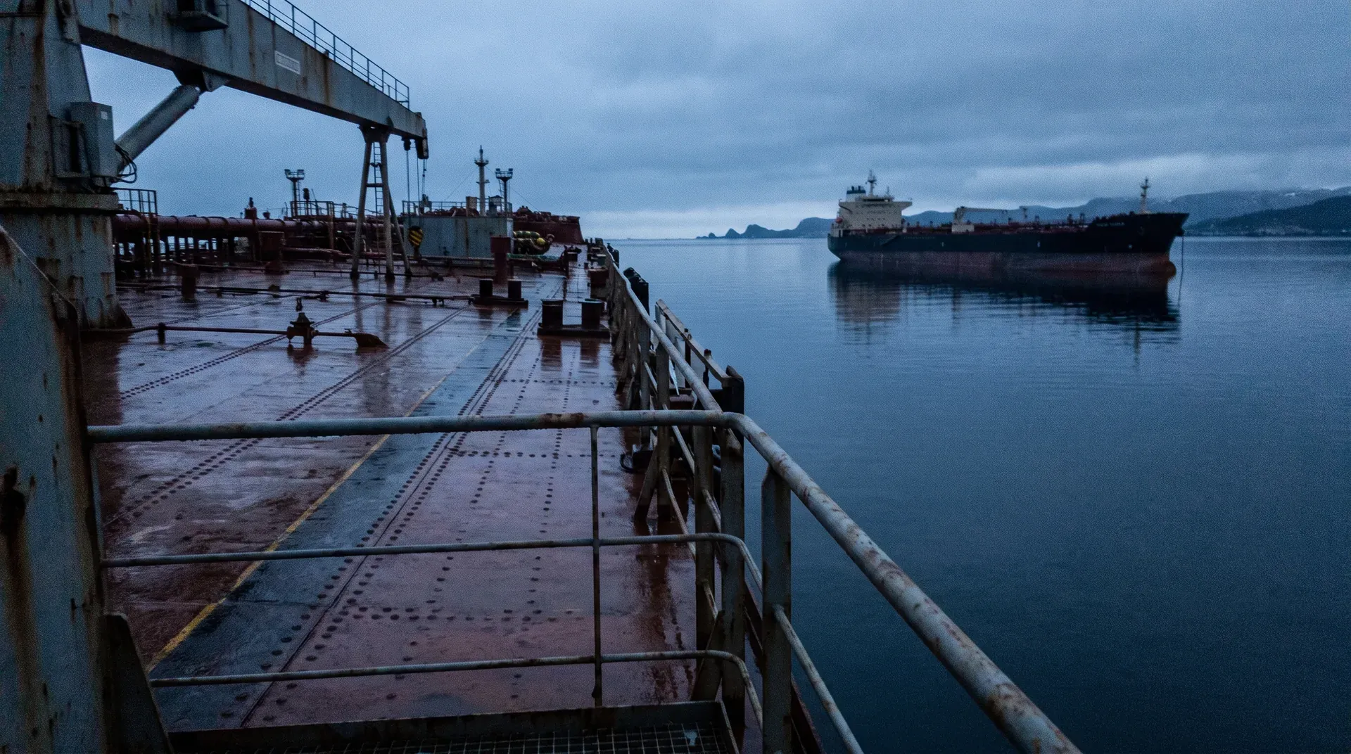 Olietanker op kalme zee gezien vanaf dek tankerschip, industriële kranen en bergen op achtergrond
