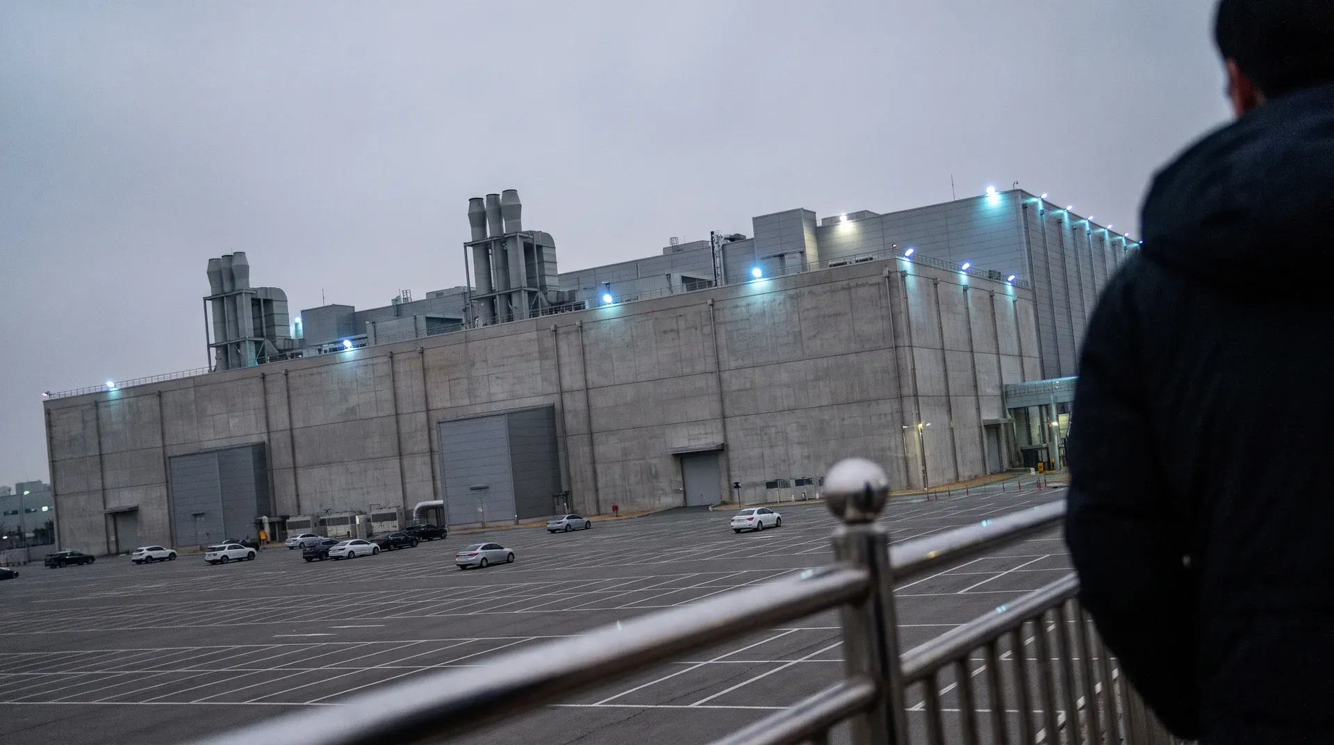 Groot industrieel gebouw met blauwe nachtverlichting, parkeerterrein met auto's, schemering