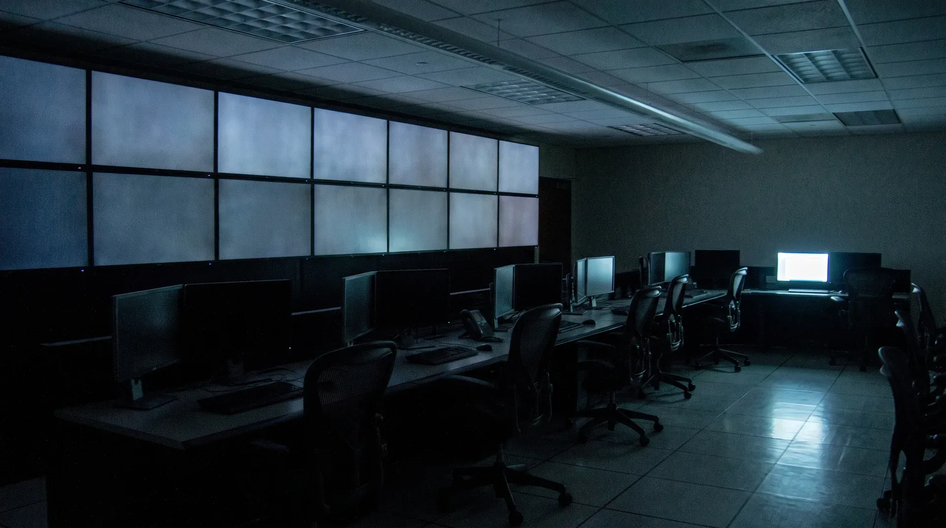 Lege computerruimte met zwakke verlichting, rijen monitoren op bureaus en zwarte stoelen.