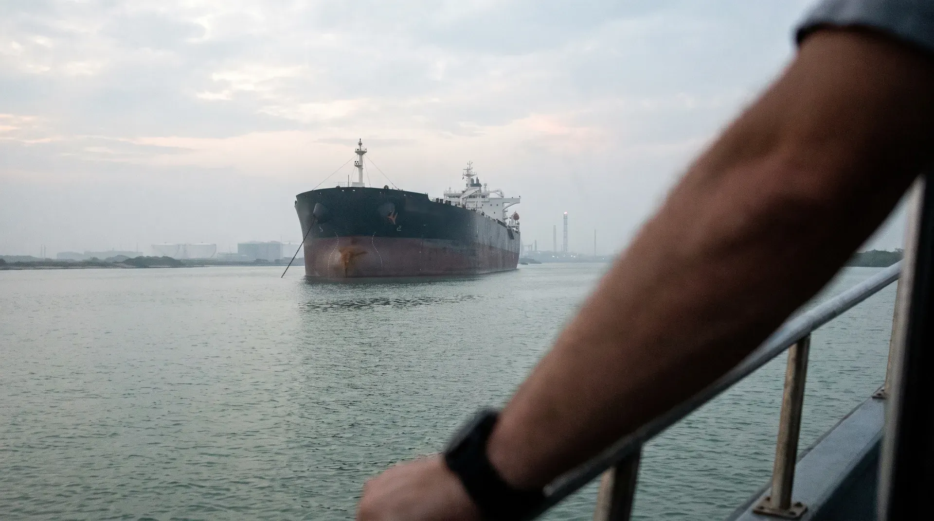 Groot olietankschip in haven, gezien vanaf dek van ander vaartuig met persoon op voorgrond