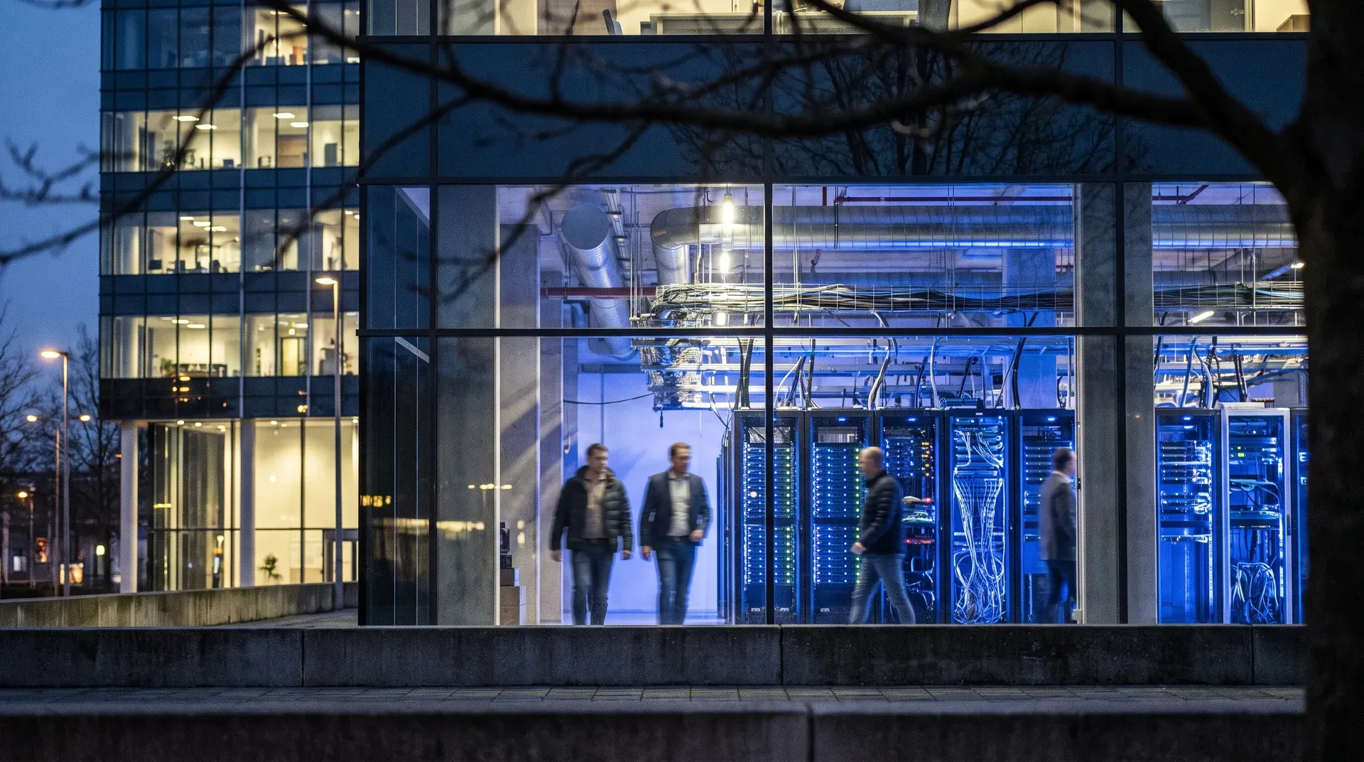 Moderne kantoorgebouw met grote glasruiten waarin serverkasten en werknemers zichtbaar zijn in blauw licht.