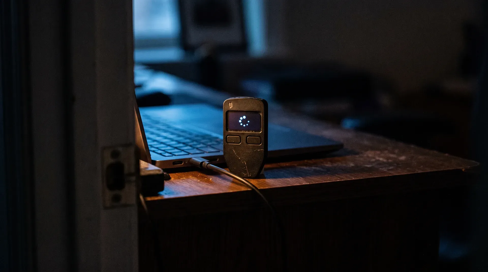 Gesloten hardware wallet met display op houten bureau in donkere kamer, laptop op achtergrond
