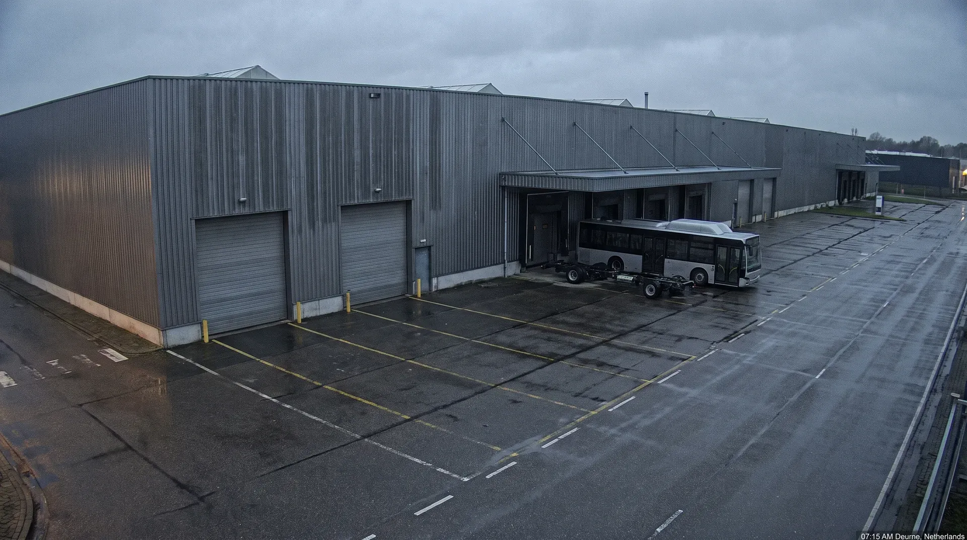Industrieel bedrijfscomplex met grijze golfplaten gebouw, roldeuren en parkeerterrein met elektrische bus.