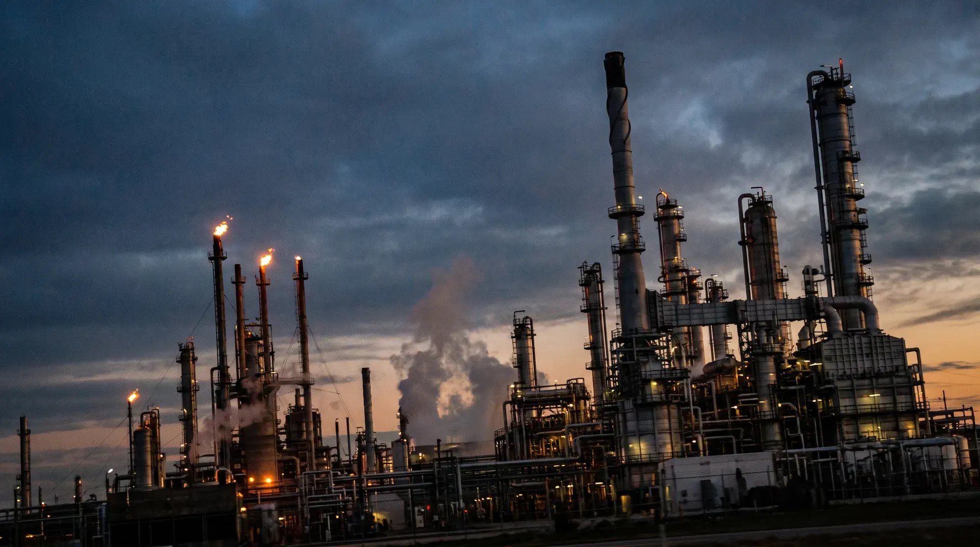 Petrochemische raffinaderij bij schemering met verlichte torens, buizen en brandende flares tegen bewolkte hemel.