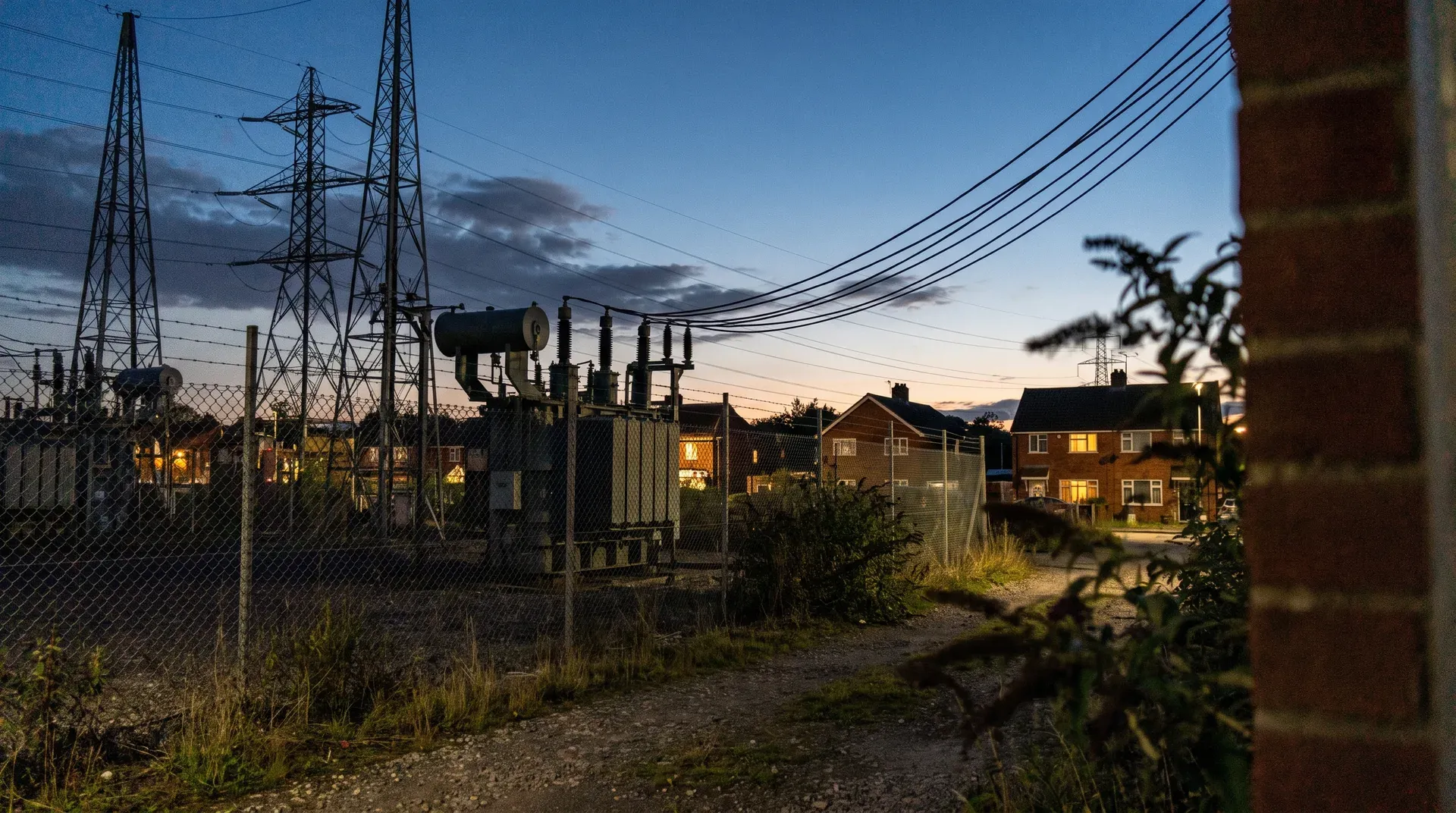 Elektriciteitsinfrastructuur met hoogspanningstorens en transformatorhuisje naast woonwijk bij schemering