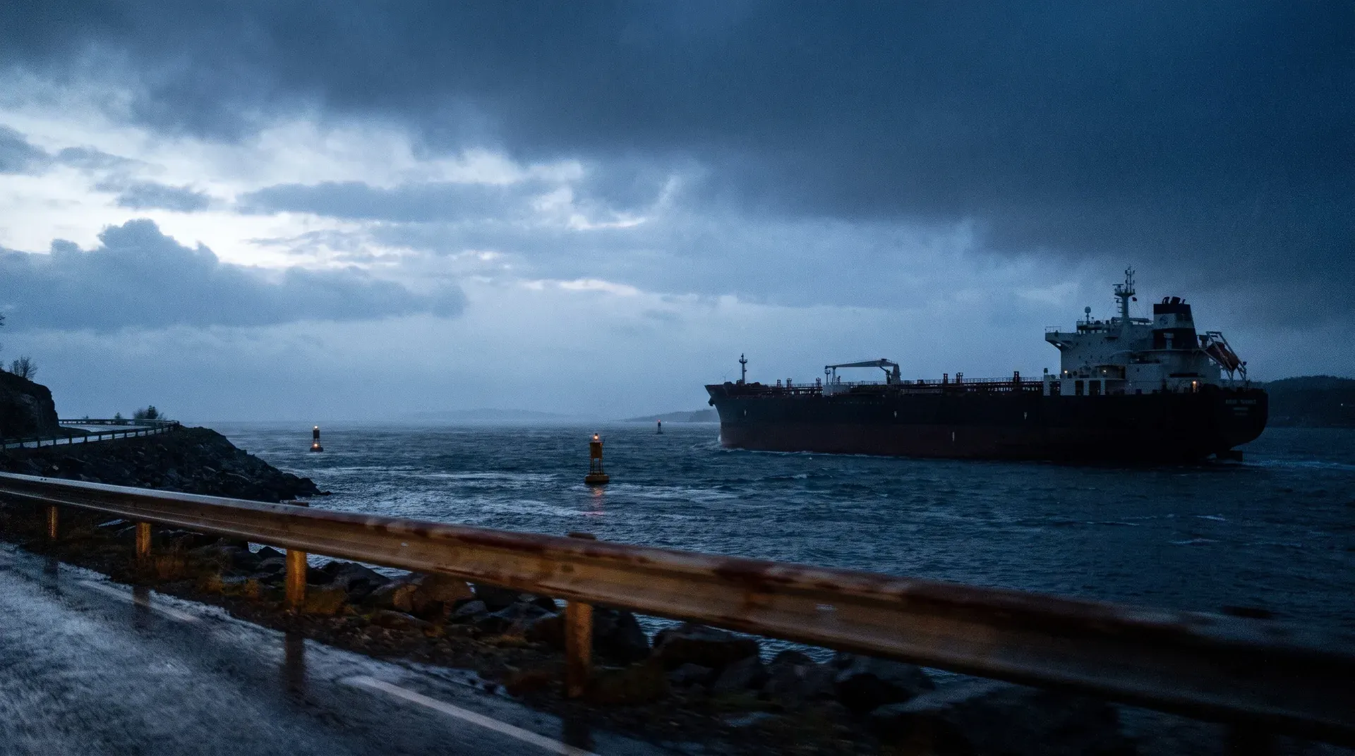 Grote olietanker voor kust bij schemering, met navigatieboeken en industriële haven in de baai.