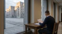 Man in donkerblauw pak zit aan houten bureau met documenten, uitzicht op moderne stenen plein met kantoorgebouwen.