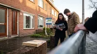 Makelaar toont brochure aan stel bij woning met TE KOOP-bord aan gevel.
