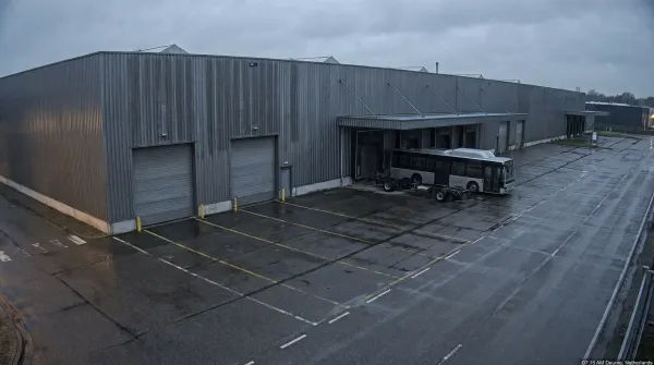 Industrieel bedrijfscomplex met grijze golfplaten gebouw, roldeuren en parkeerterrein met elektrische bus.
