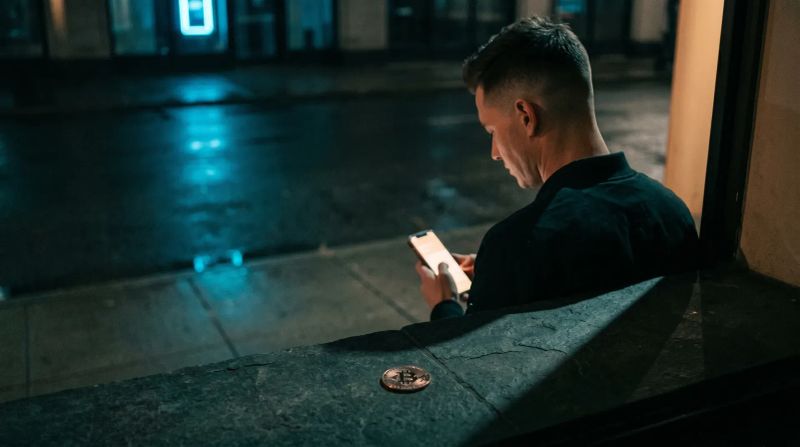 Man in donkere kleding zit 's nachts op stoep, smartphone in handen, Bitcoin-munt op grond
