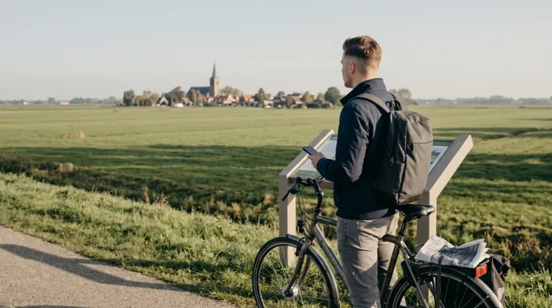 Man met fiets en kaart kijkt uit over groen landschap met dorp in de verte.