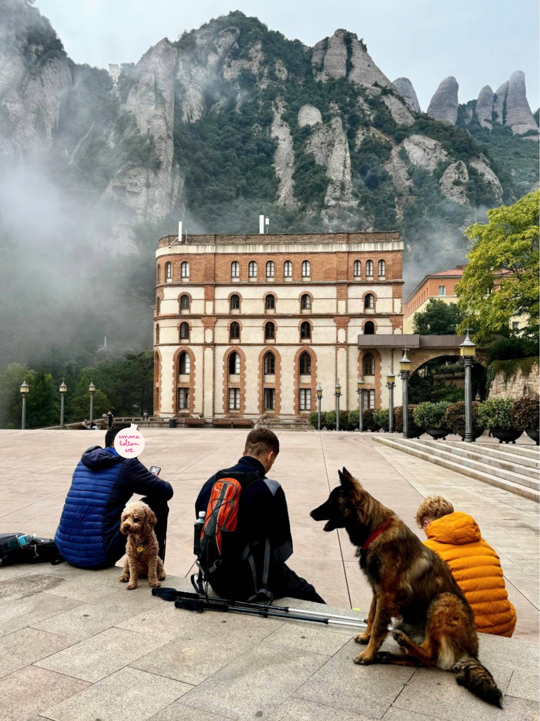 Three tourists and two dogs sit in the foreground with their backs to the camera. Across the empty square is a yellow building at Montserrat, with the monoliths in the background. It is slightly foggy and the people are wearing puffer jackets. 