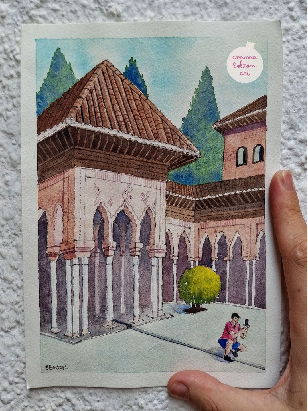 Watercolour painting of a man in a red shirt crouched down to take a photo. Behind him is the pink building filled with beautiful archways in the Patio de los Leones at the Alhambra palace in Granada.