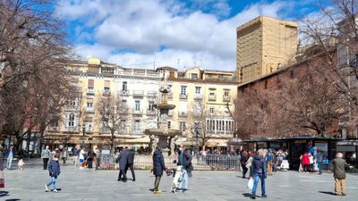 People walking across a large, busy square surrounded by naked trees and pastel-coloured apartment blocks. A fountain is featured in the centre and it's a sunny day.
