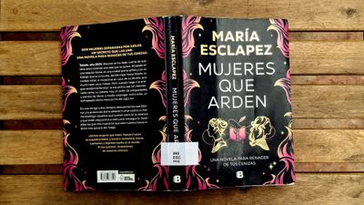 A copy of the book Mujeres que arden by María Esclapez on a wooden background. The book has a black cover with gold and pink details.