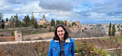 A white woman wearing glasses and a denim jacket is smiling at the camera. In the distance is the Alhambra palace and Granada.