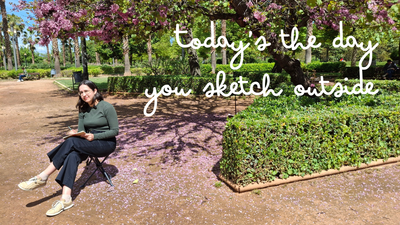 Today's the day you sketch outside. A white woman in a green top and black jeans is sketching in a park under a judas tree. 