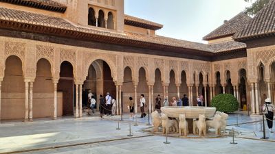 The grey stone fountain comprising a bowl held up on the backs of stylised lions. Behind the fountain, a line of tourists is walking into the Alhambra palace. 