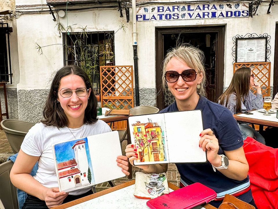Two white women sitting in a coffee shop in the Albaicín in Granada and holding up watercolour sketches with big smiles.