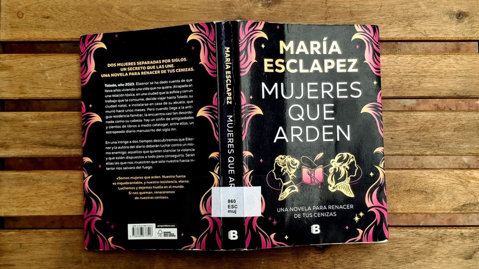 A copy of the book Mujeres que arden by María Esclapez on a wooden background. The book has a black cover with gold and pink details.