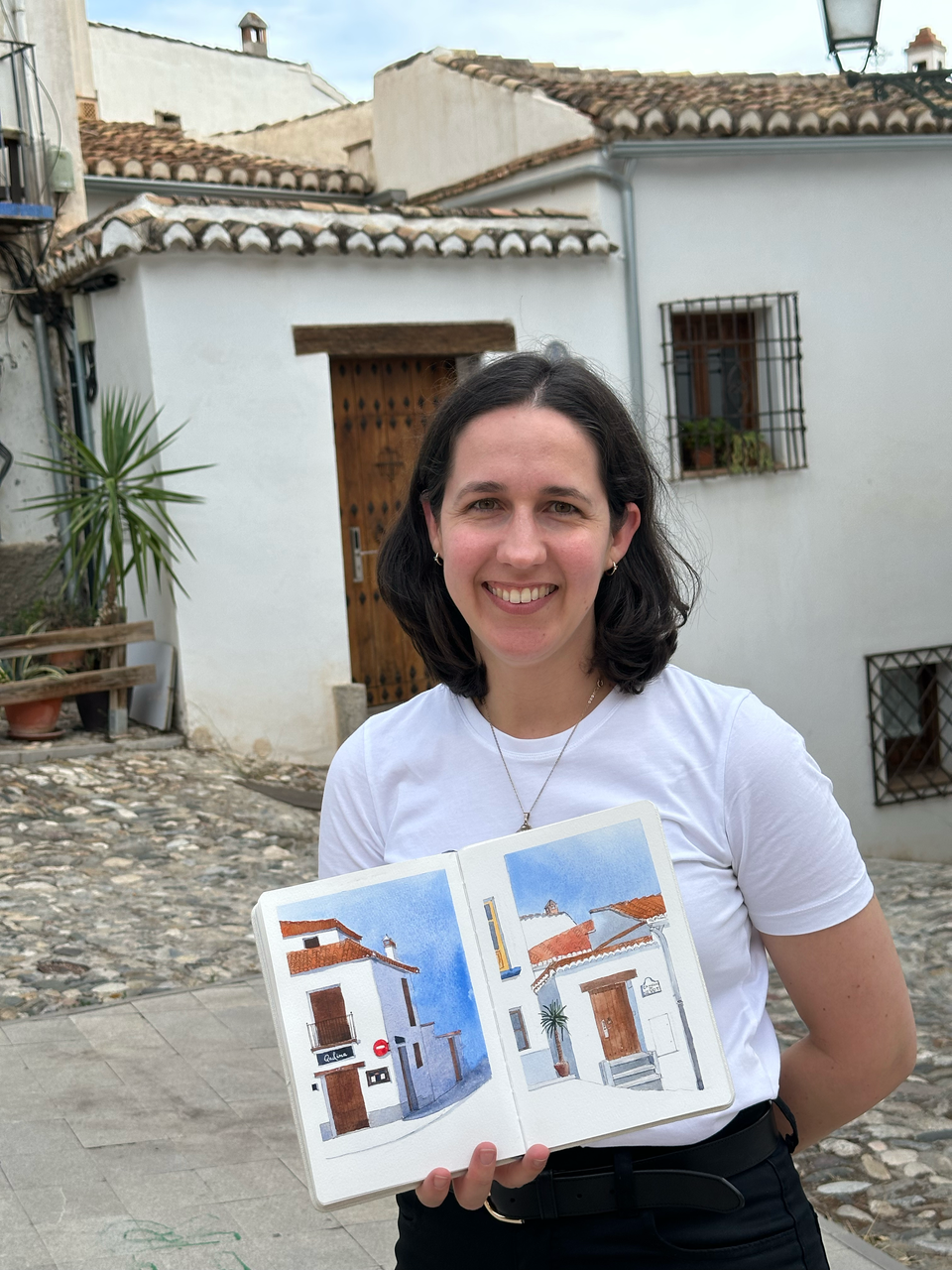 A white woman with brown hair and wearing a white tshirt is smiling at the camera. She's holding up her watercolor sketchbook of white buildings in the Albaicín (which are behind her). 