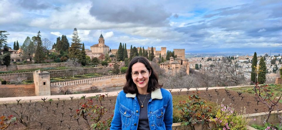 A white woman wearing glasses and a denim jacket is smiling at the camera. In the distance is the Alhambra palace and Granada.