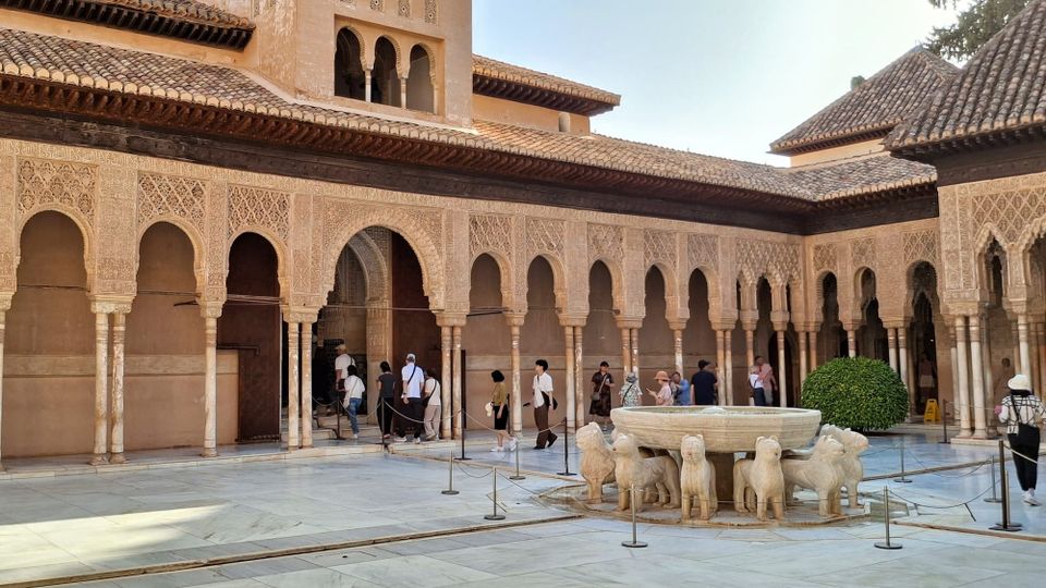 The grey stone fountain comprising a bowl held up on the backs of stylised lions. Behind the fountain, a line of tourists is walking into the Alhambra palace. 