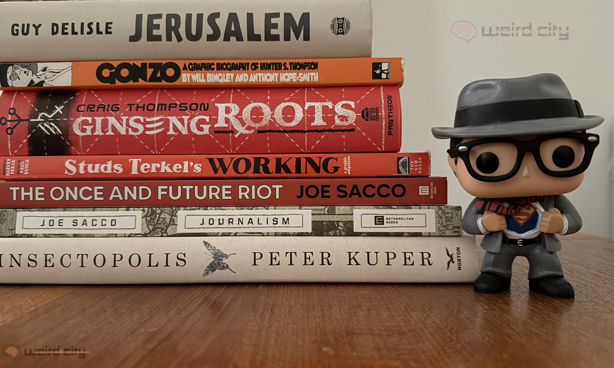 A stack of graphic novels that include Jerusalem, Gonzo: A Graphic Biography of Hunter S. Thompson, Ginseng Roots, Studs Terkel's Working, The Once and Future Riot, Journalism, and Insectopolis. Beside the stack is a Clark Kent Funko Pop figure. 