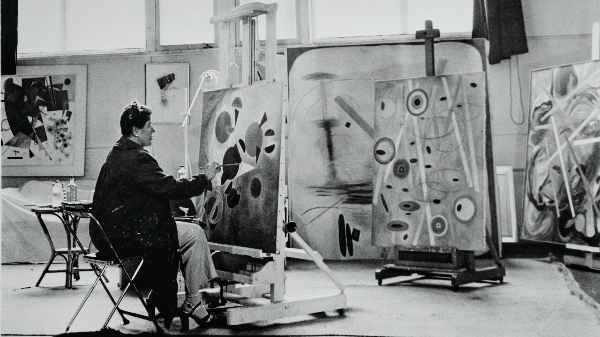 Woman at left painting abstract forms on a canvas in a studio with other canvasses.
