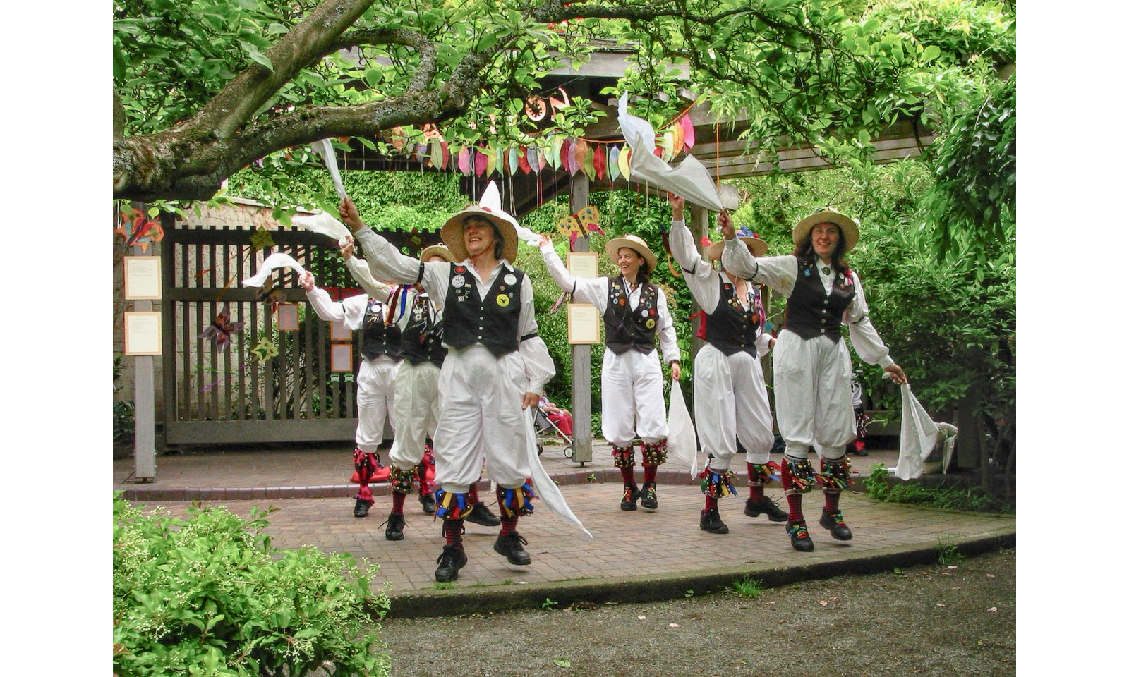 Photo of 6 Morris dancers on a small stage in a park, wearing costumes and holding white handkerchiefs to their right
