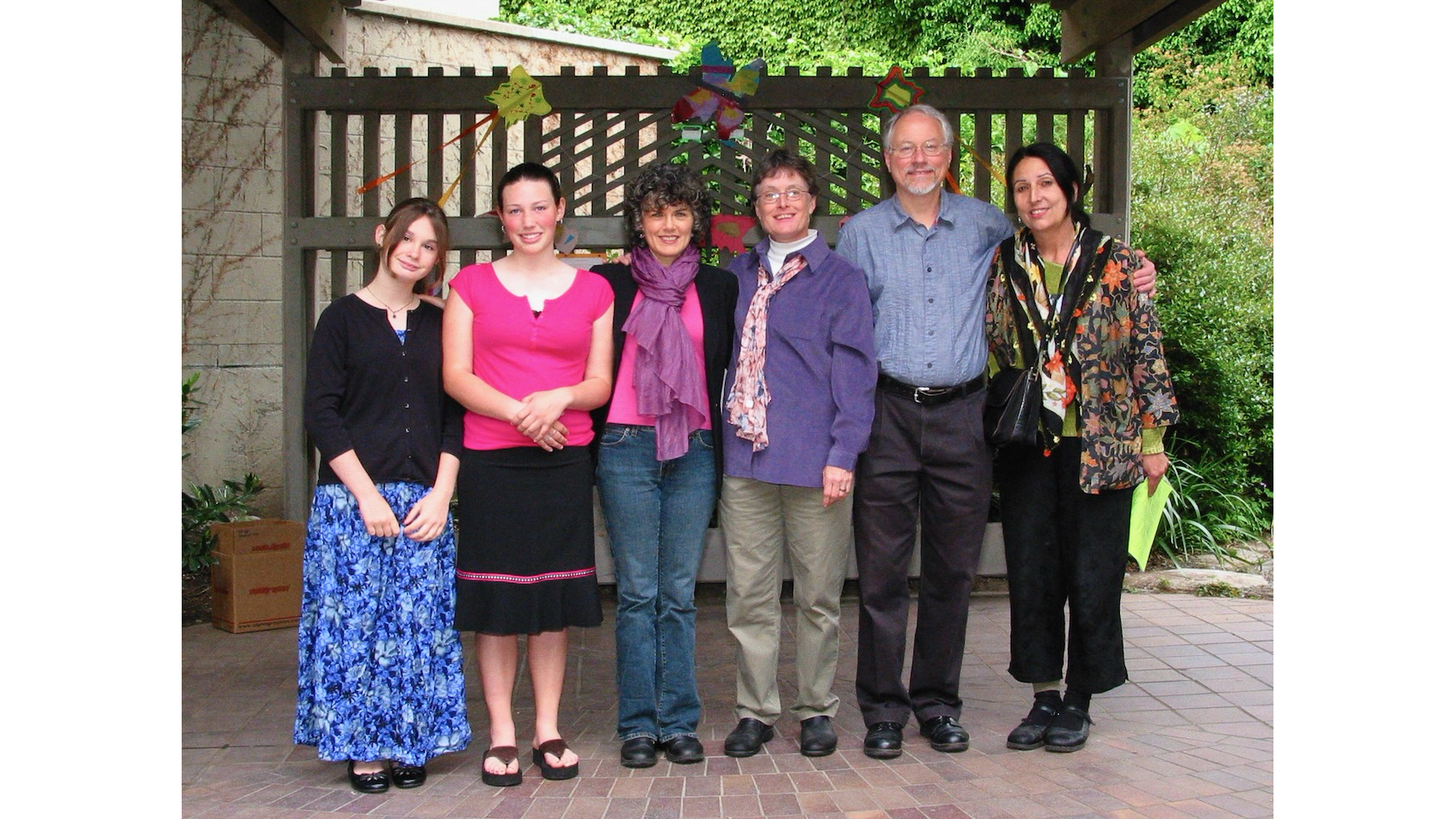 Photo of 2 teen girs and4adults in a row, smiling, on a small stage in a park with a wooden slat background