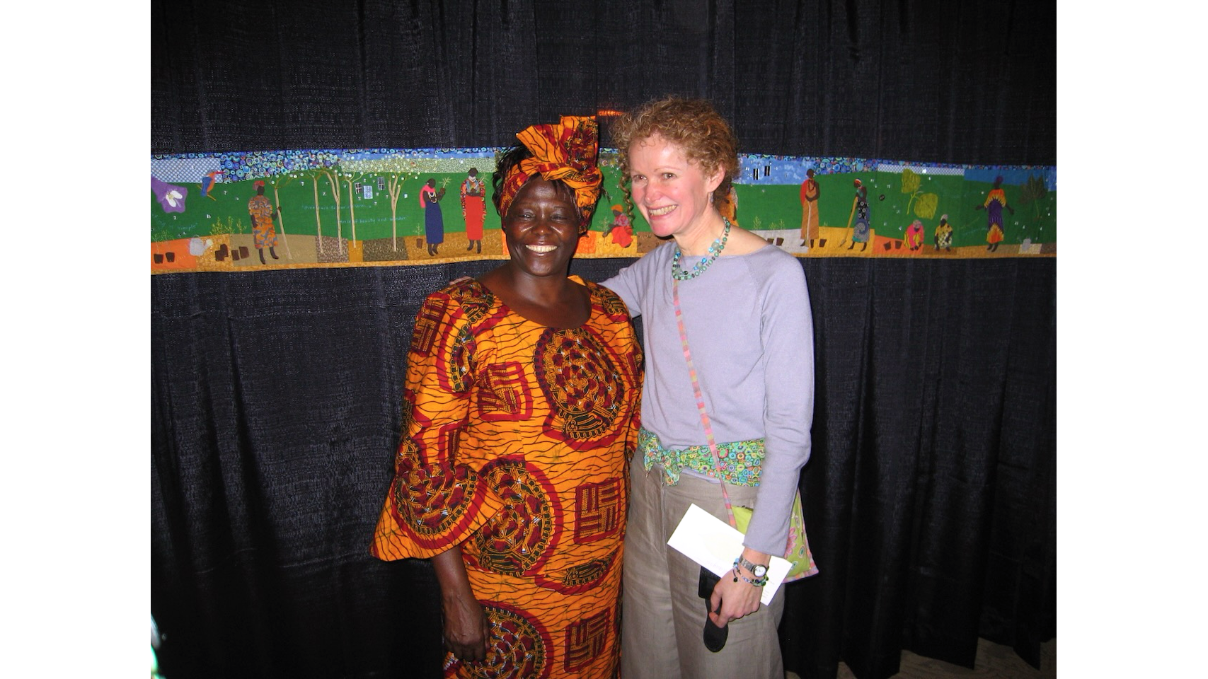 Photo of African woman in orange print dress and matching headband next to woman in light blue top, both smiling at camera