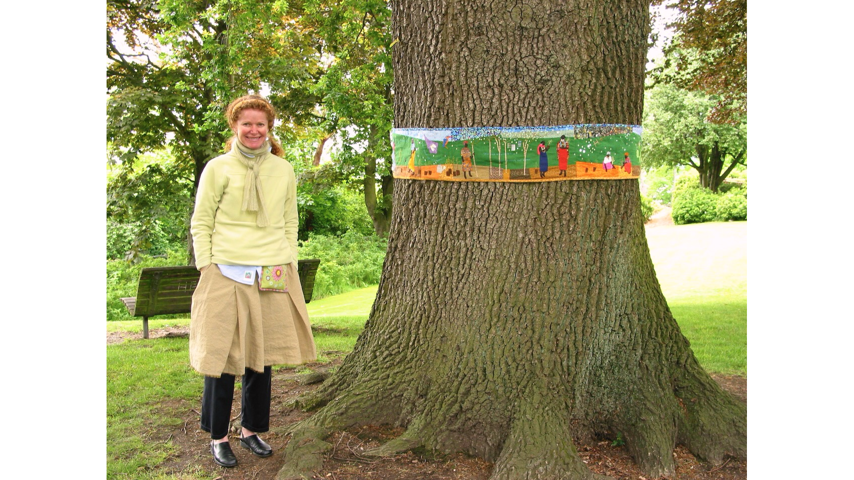 Photo of a woman in yellow sweater and bron skirt, smiling, next to a tree trunk with an embroidered green belt around it.