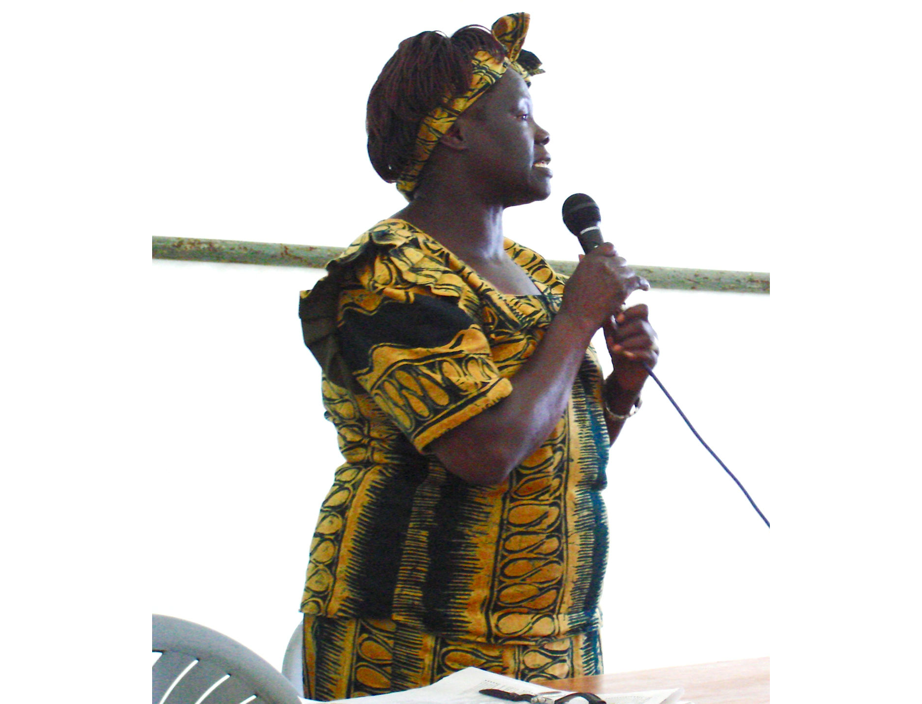 Photo of Wangari Maathai looking to her left, wearing a yellow print dress and matching headband, holding a microphone and speaking.