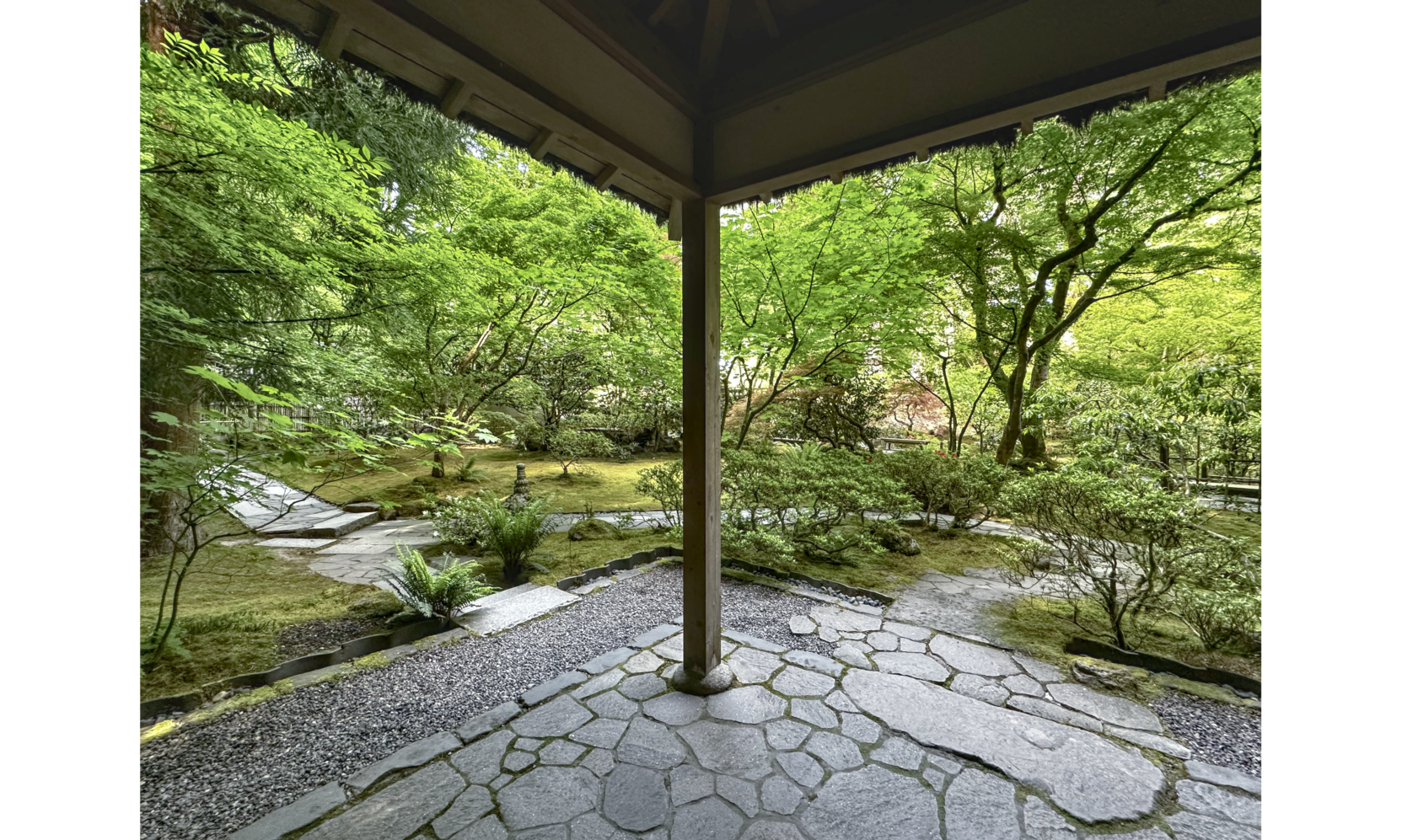 Photo looking out from an open veranda with stonework floor and wooded post and roof, onto garden with stone paths and trees and shrubs
