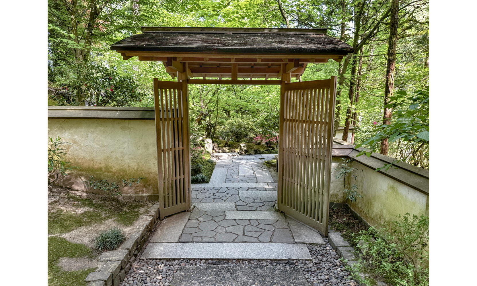 Photo of blocked stone path leading through open wooded gate with long horizontal roof, connected to two stone walls, and trees in background