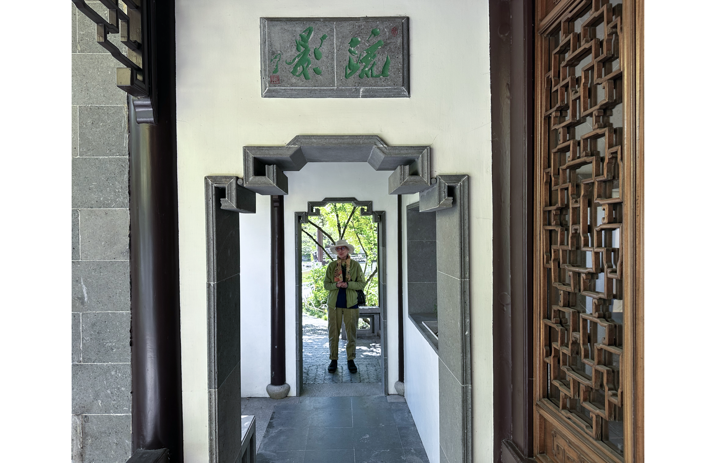 Photo of Sandra in green jacket and hat in background arch, with a second arch, carved window covering, and two stone wall hanging with green lettering in foreground