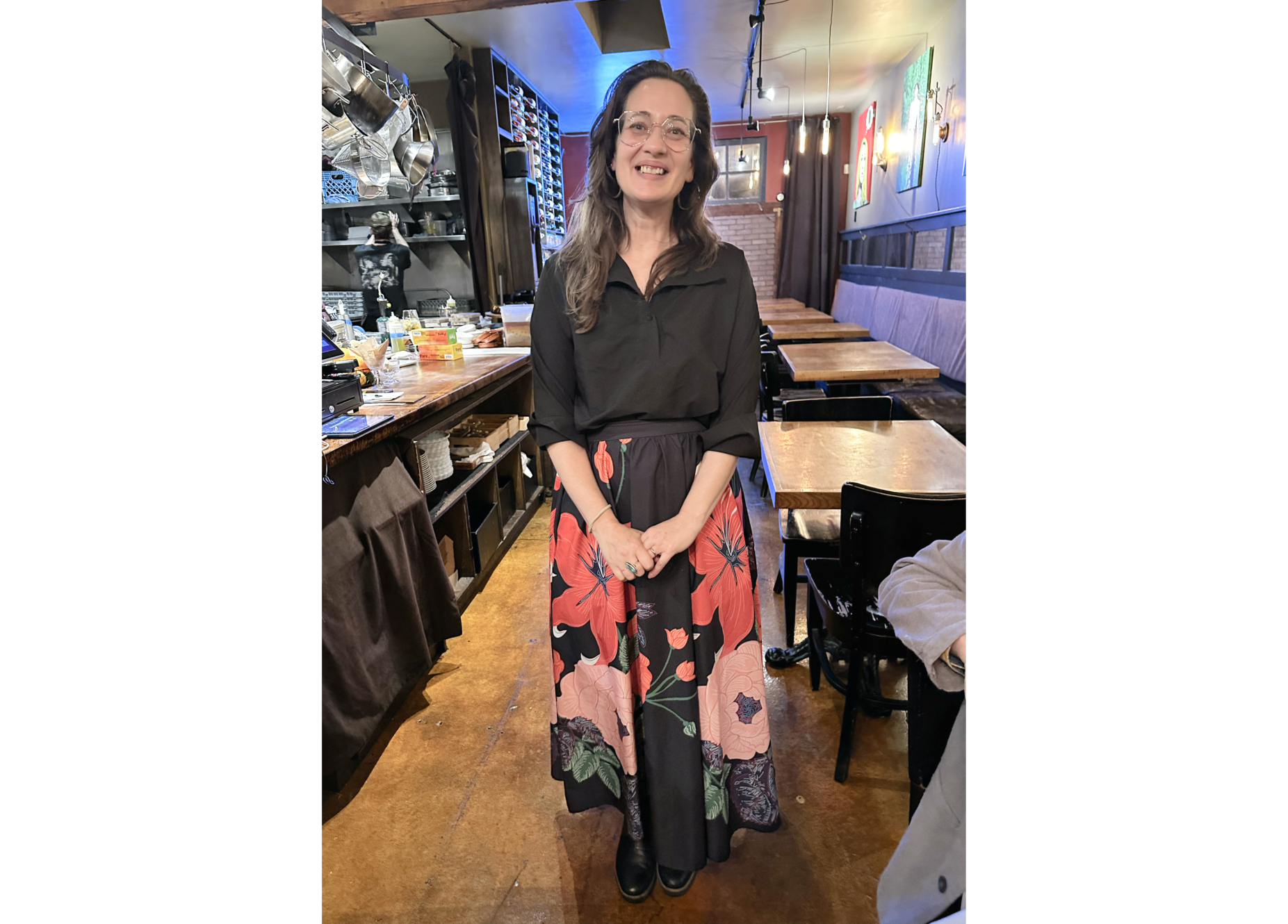 Phot of woman in classes, black blouse and large red flower-patterned long skirt, with tables in background