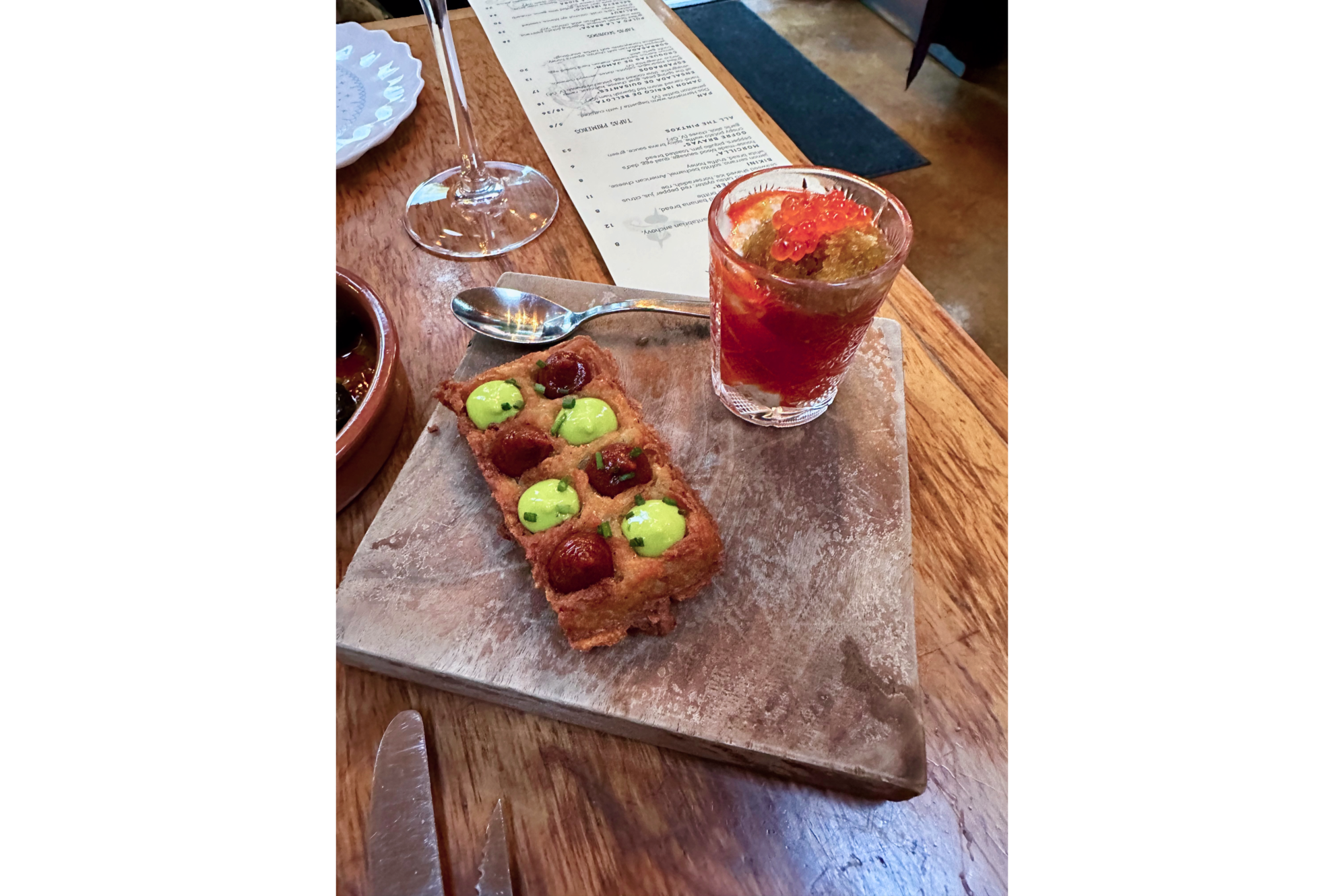 Photo of section of dining table with glass of red food and waffle with 8 small servings of green and brown, on wooden tray