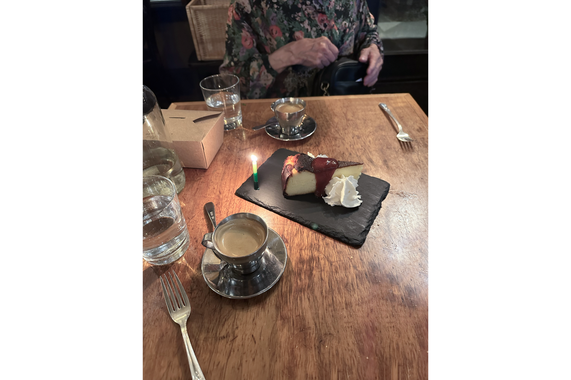 Photo of dining table with two silver cups of espresso and tray of cheesecake with red quince past and whipped cream, next to a lit green candle