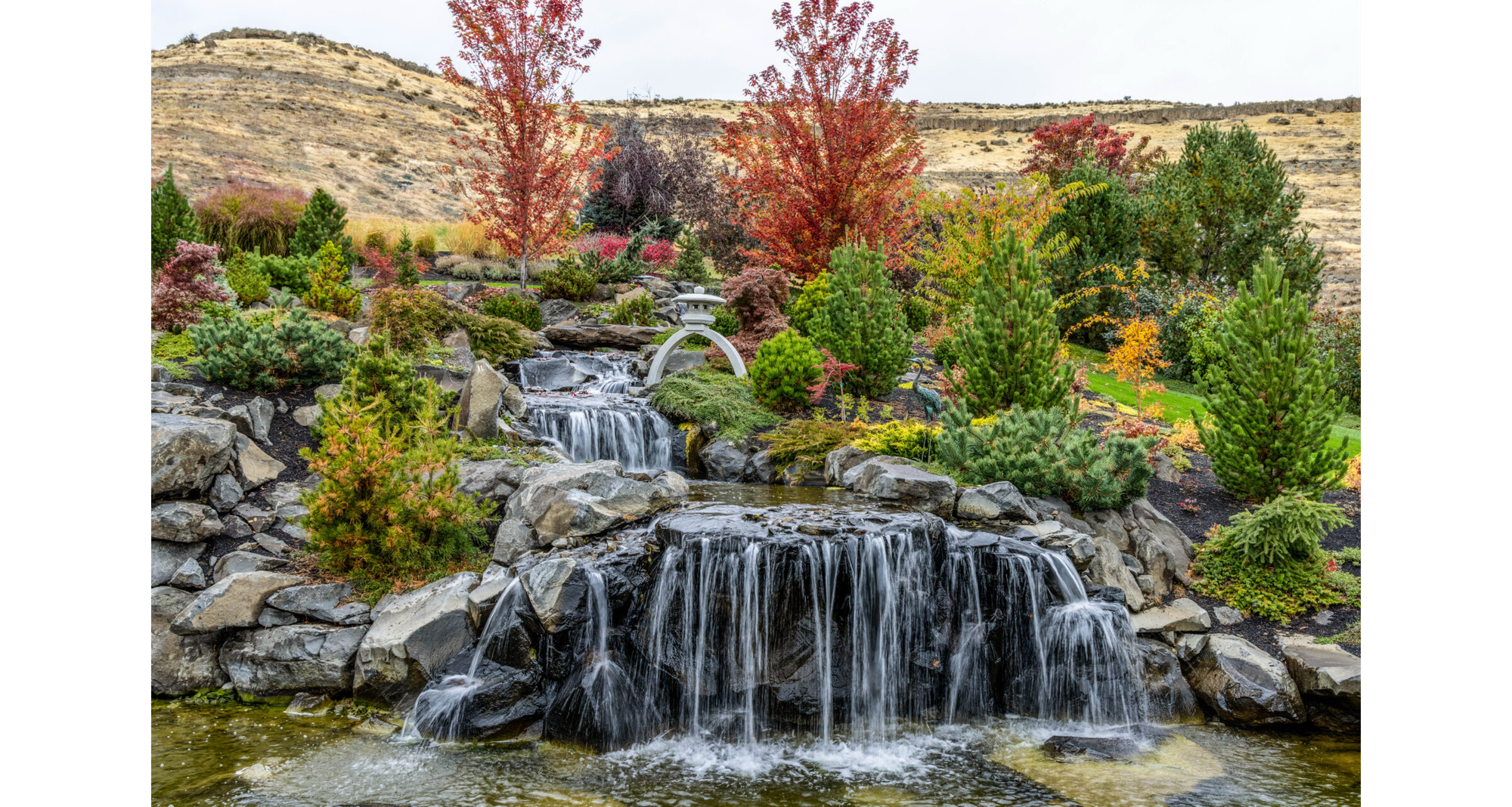 Photo of water flowing over multilevel orck formations, with green, gold, a nd red shrubs, flowers, and tress surrounding it, and a white Japanese-style sculpture in center, with brown hills in background