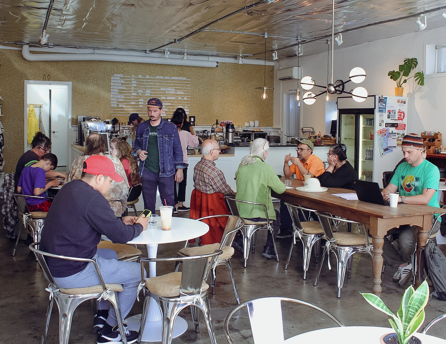Photo of the interior of a coffehouse, w ith people sitting at long tables conversing or reading, and baristas in the background behind a counter.